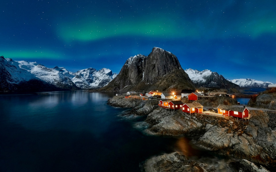 Nordlichter über roten Fischerhütten in einem norwegischen Fjord, umgeben von schneebedeckten Bergen bei Nacht.